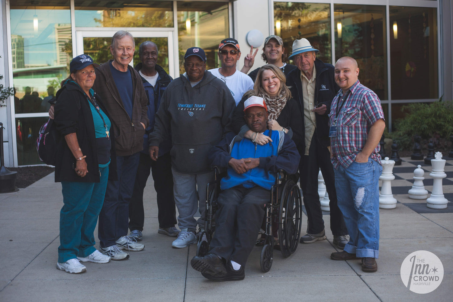 Group of people standing and smiling outside a building, with one person seated in a wheelchair in the front. The atmosphere reflects community and support, representing the work of Room In The Inn in Nashville.