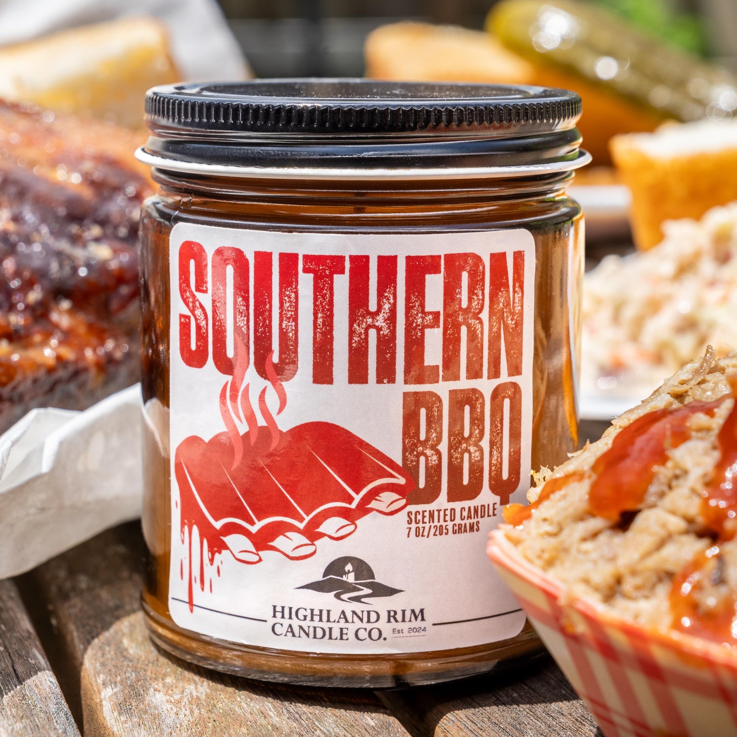 Southern BBQ candle by Highland Rim Candle Co. on a picnic table surrounded by barbecue food, celebrating Southern hospitality and flavor.