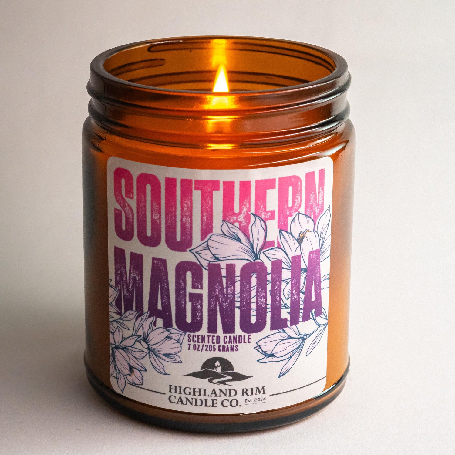 Amber glass Southern Magnolia candle by Highland Rim Candle Co., lit in a studio setting, emitting a soft glow with a purple magnolia flower label design.