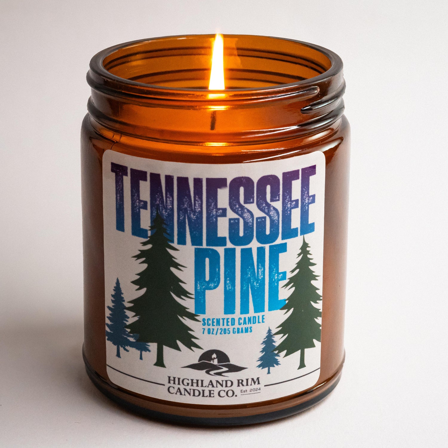 Amber glass Tennessee Pine candle by Highland Rim Candle Co., lit in a studio setting, with a green and blue label design featuring pine trees.