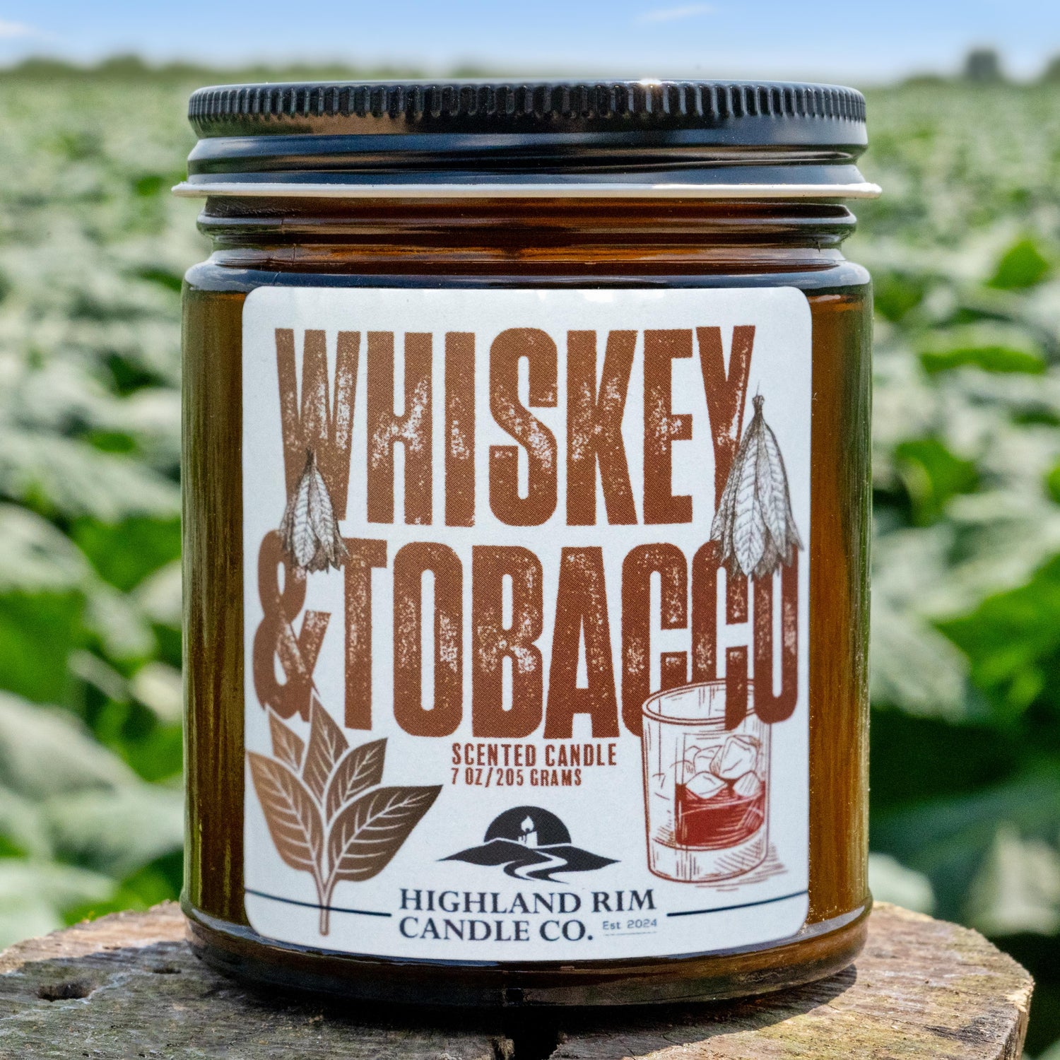 Highland Rim Candle Co.'s Whiskey & Tobacco scented candle in amber jar, displayed with fresh tobacco leaves in the background, showcasing the label's vintage whiskey and tobacco motif.